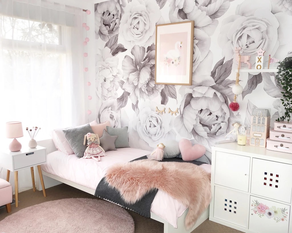 : اتاق دختر بچه با دکوراسیون شیک و مدرن که تخت سفید، بالش قلب و ستاره، کاغذ دیواری گل دار، روتختی صورتی و تابلو دکوری قو دارد